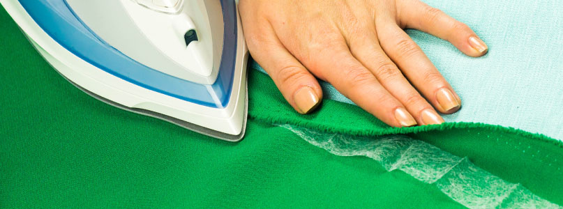 Feminine hand using an iron to apply basting tape or heat bonding fabric tape to the folded over hem of green fabric.