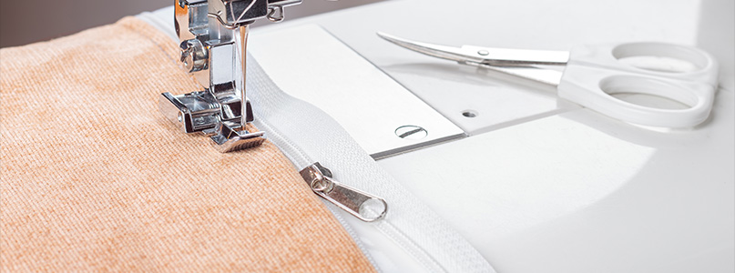 An image of Professional sewing machine securing a white zipper to textured orange upholstery fabric after being placed with double sided adhesive fabric tape.